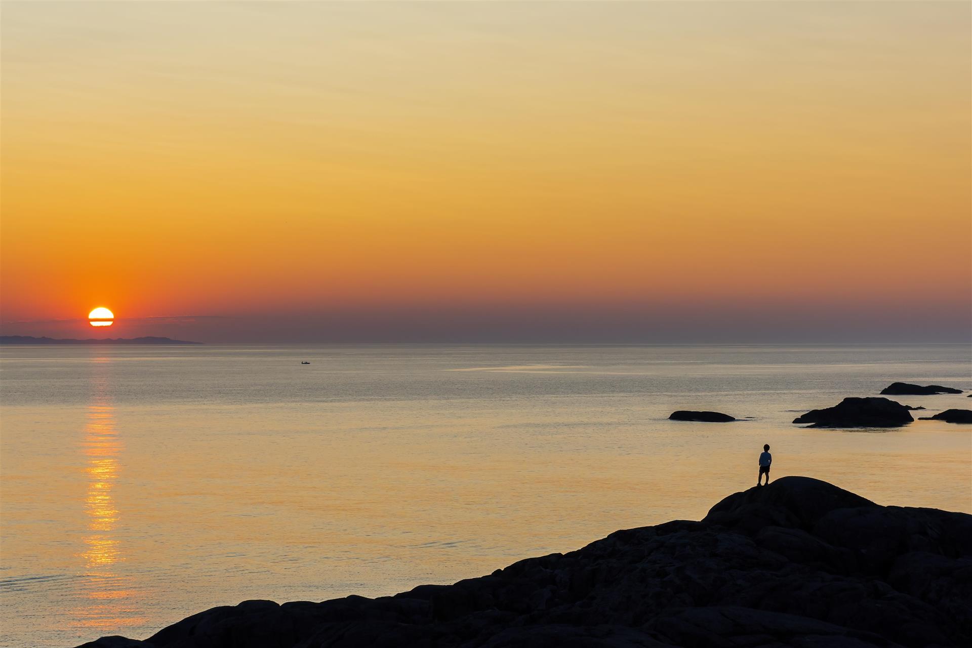 En person på et svaberg ser utover havet og en gyllen solnedgang 