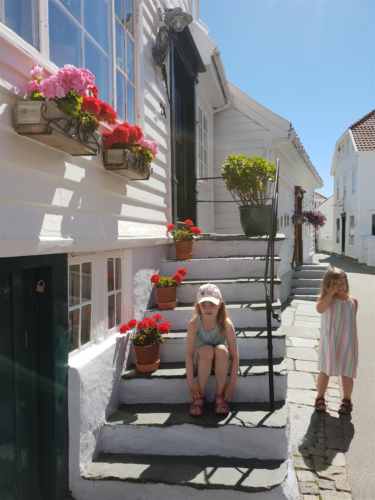 Sommerkledde jenter ved trapp blant hvitmalte trehus og blomster i gamle Skudeneshavn - Klikk for stort bilde
