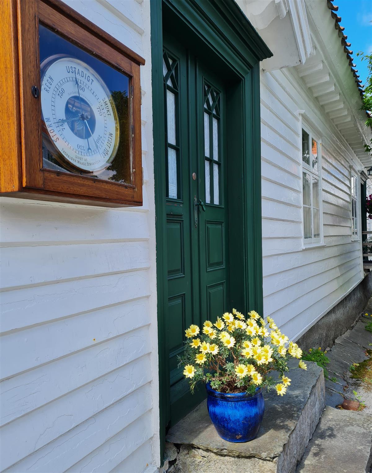 Barometer og grønn dør mot hvitmalt trehus og gule blomster i en blå krukke - Klikk for stort bilde